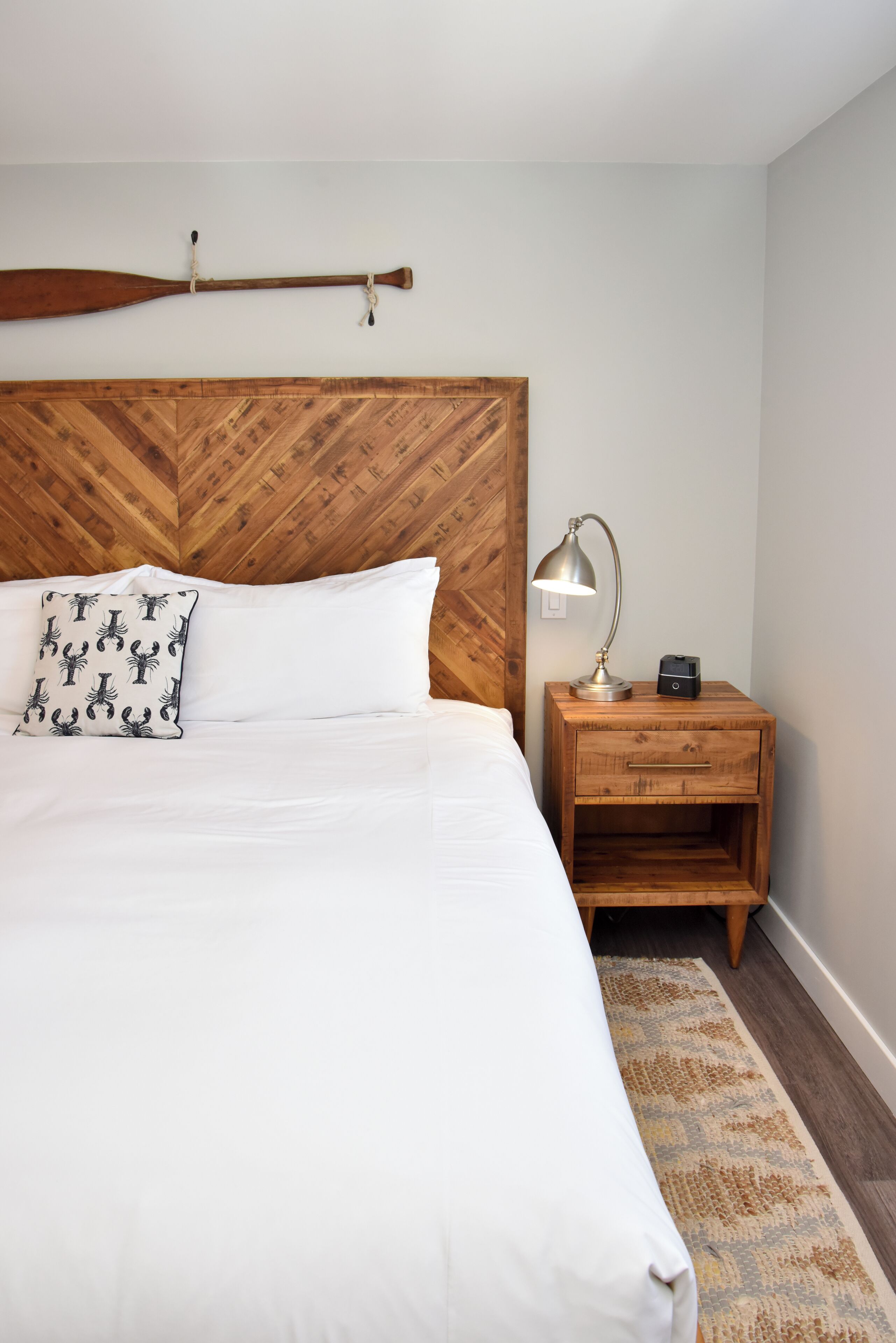 A neatly made bed with white linens and a patterned pillow, a wooden headboard, a bedside lamp, and a small wooden nightstand with a lamp and a phone.