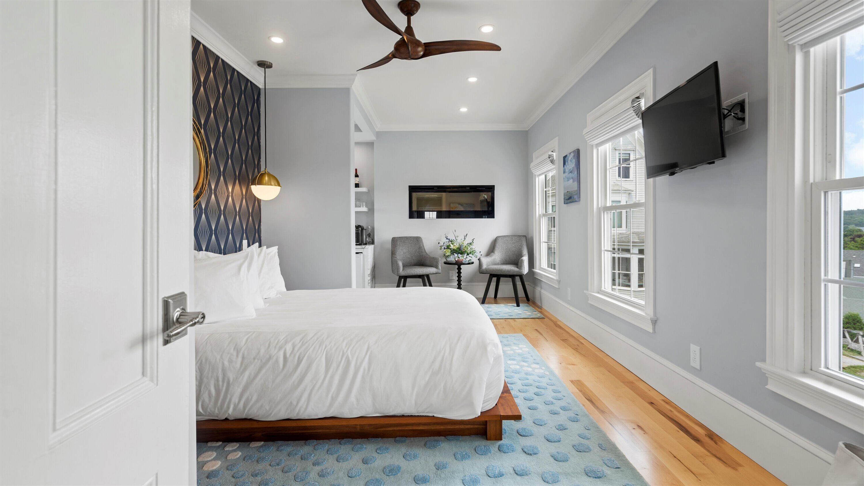 A bedroom with a bed, two chairs, a ceiling fan, a TV, and a window.