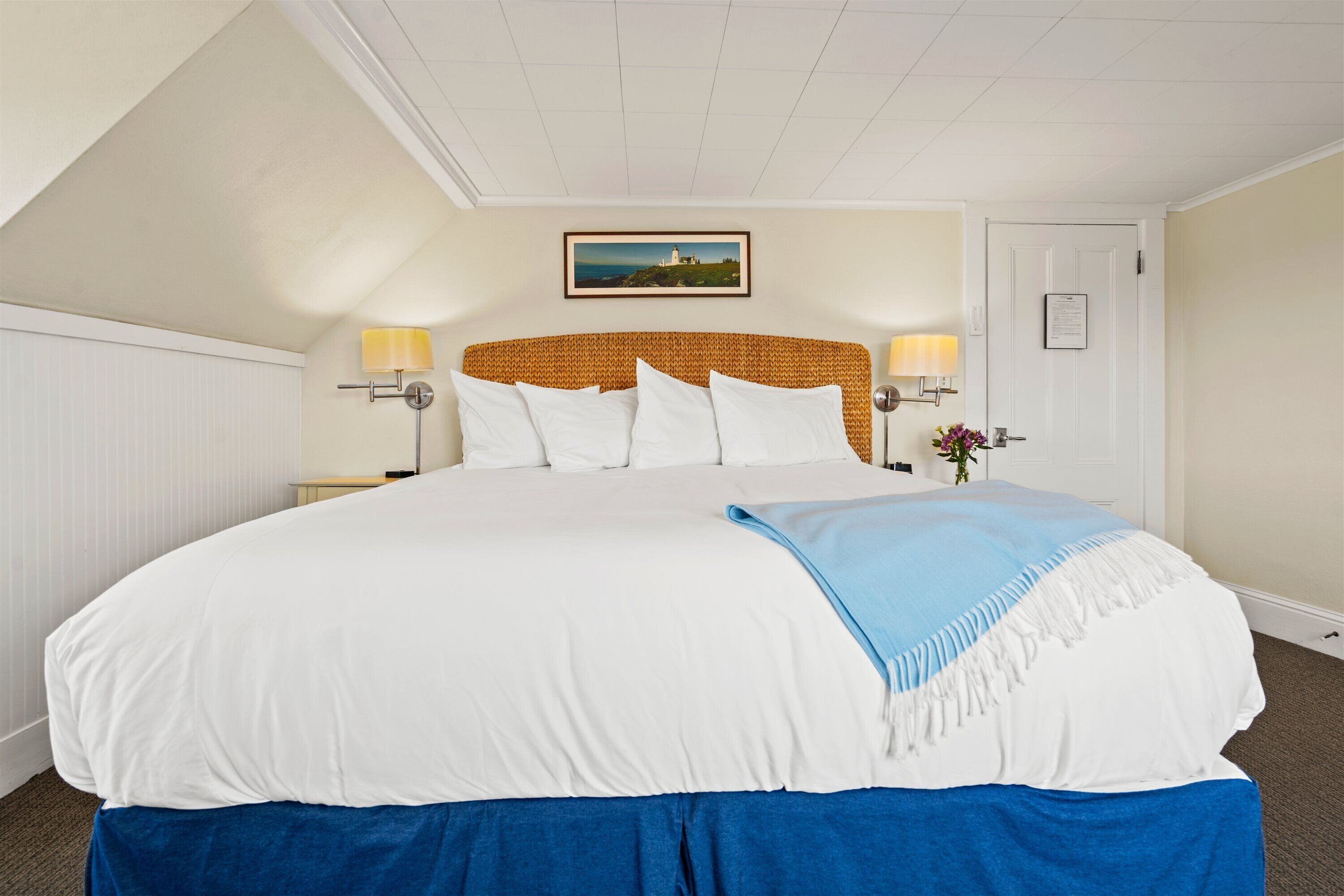 A large bed with white bedding and a blue throw blanket, flanked by two bedside lamps and a framed picture on the wall.