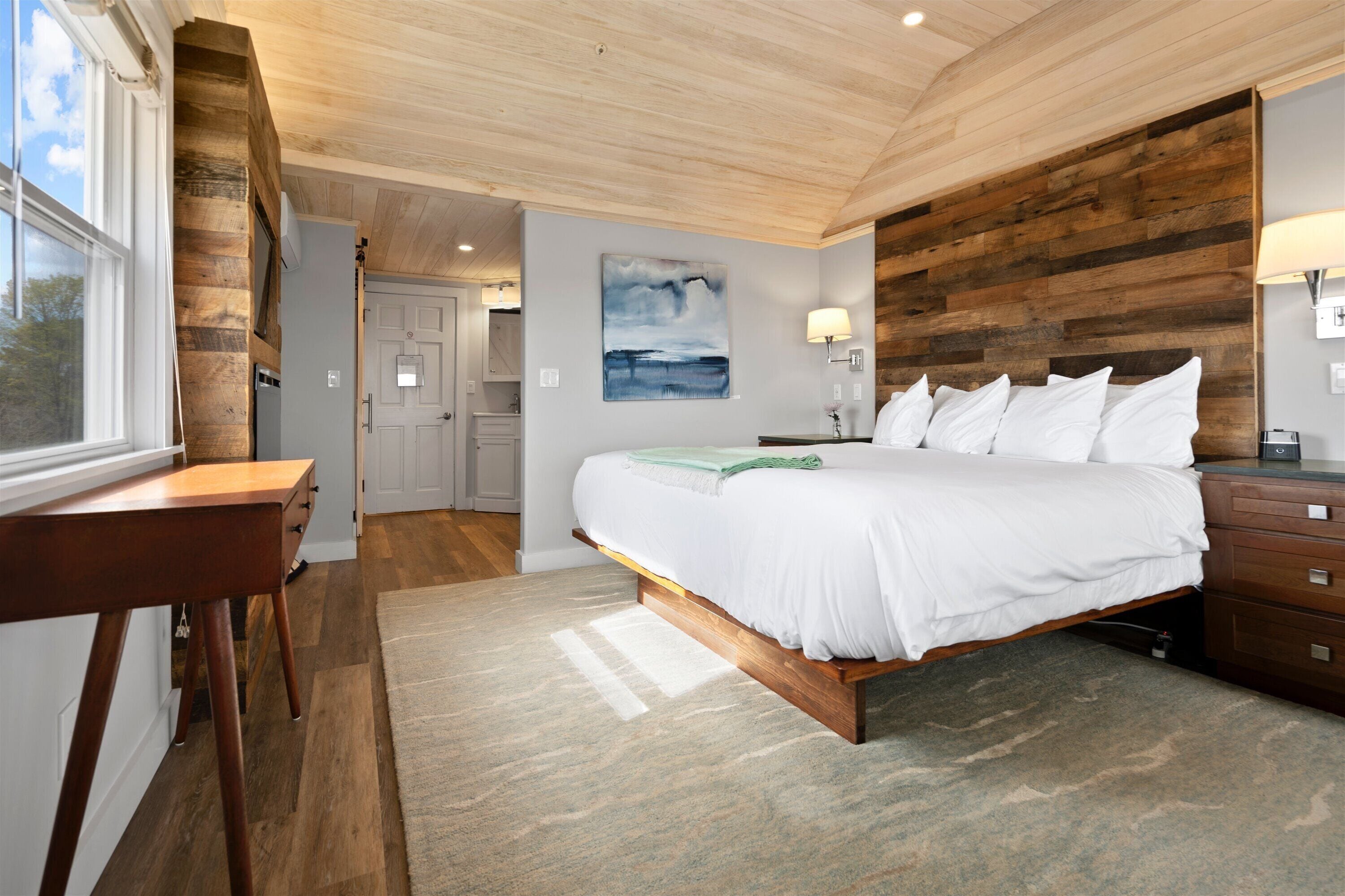 A bedroom with a wooden headboard, a bed with white linens, a wooden desk, and a view of the outdoors through a window.