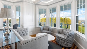 A bright, well-lit room with a glass table, a tufted sofa, patterned armchairs, and large windows offering a view of the outdoors. - Topside Inn (Boothbay Harbor)
