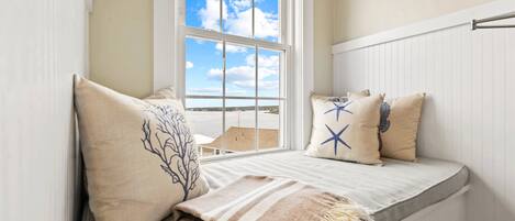 Una habitación con asiento junto a la ventana que ofrece vistas del cielo y un edificio. El asiento tiene almohadas con diseños de estrellas y árboles.
