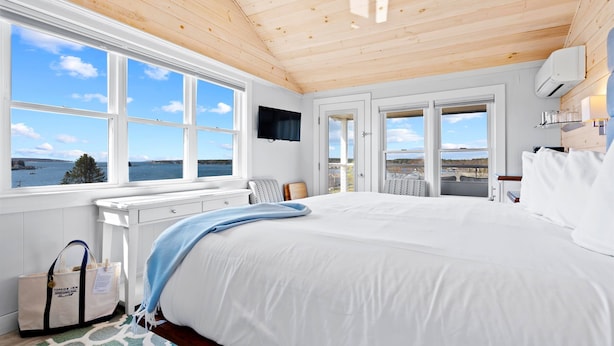 A bedroom with a large bed, white walls, wooden ceiling, and a view of the water. - Topside Inn (Boothbay Harbor)