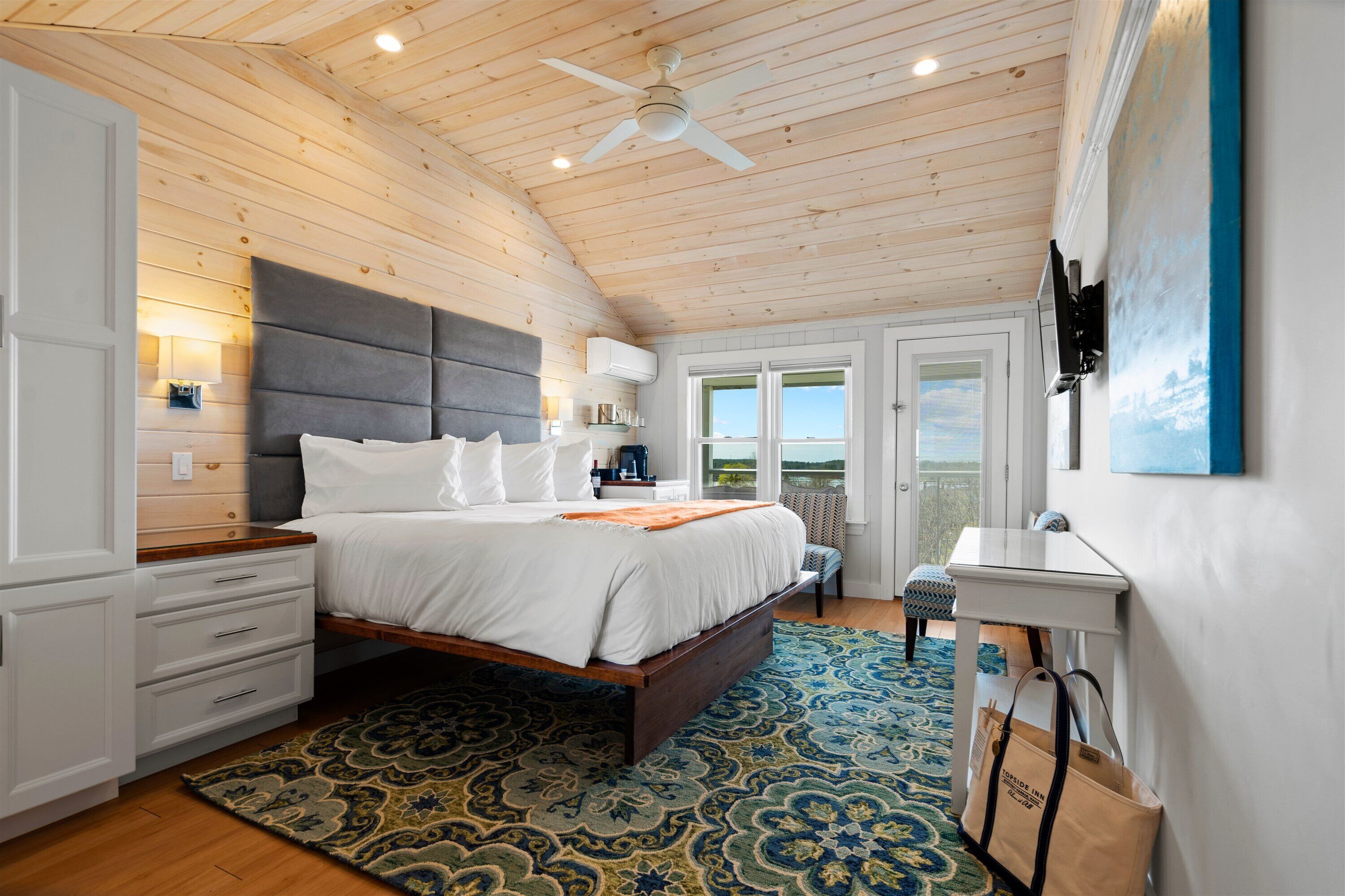 A bedroom with a wooden ceiling, a bed with white bedding, a nightstand, a desk, and a view of the outdoors through a door.