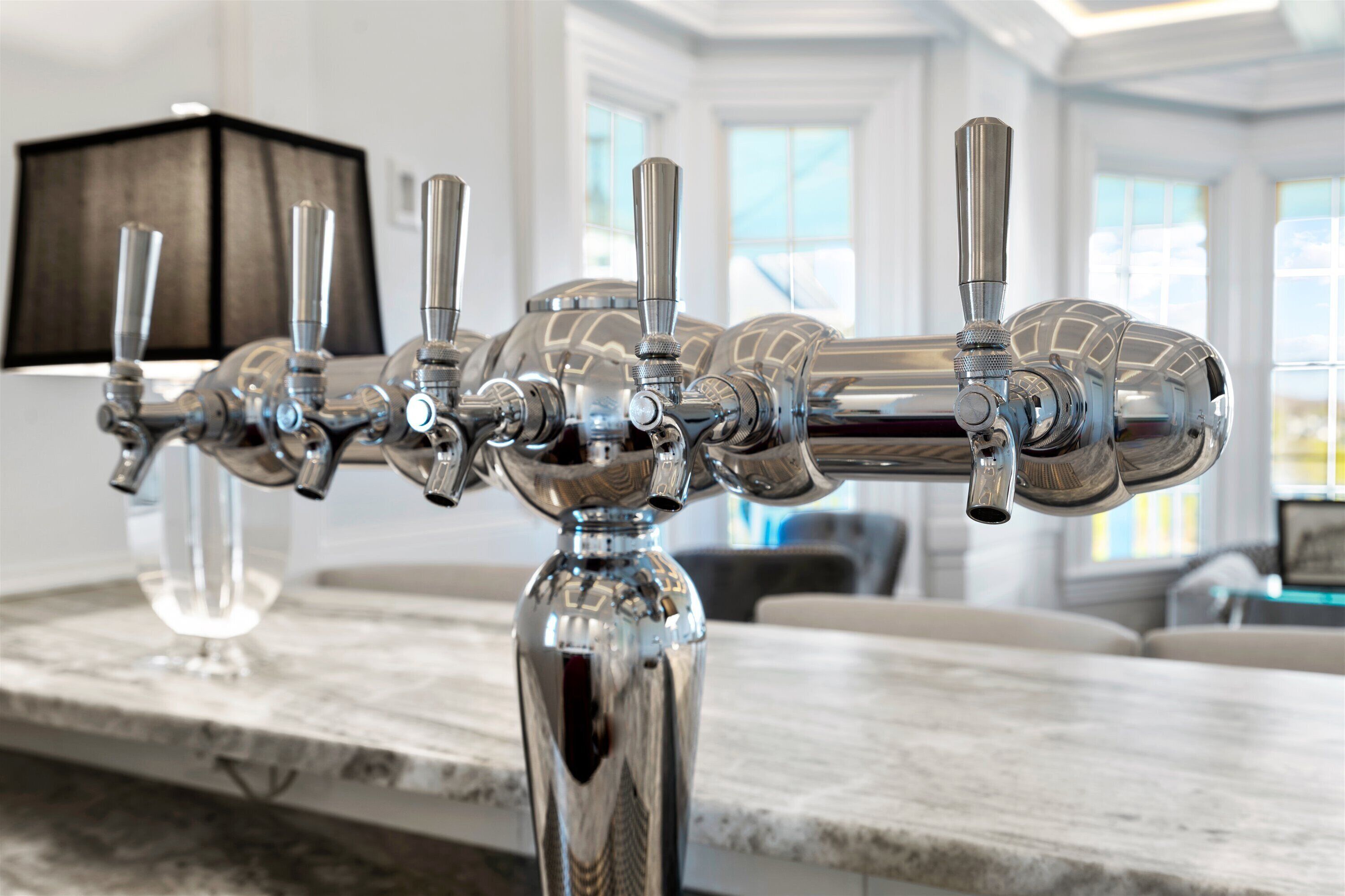 A row of stainless steel bar faucets with multiple spouts and handles, mounted on a marble countertop in a well-lit room with large windows.