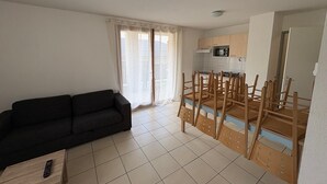 Una sala de estar con un sofá negro, una mesa de comedor con sillas, una zona de cocina y un ventanal corredero.