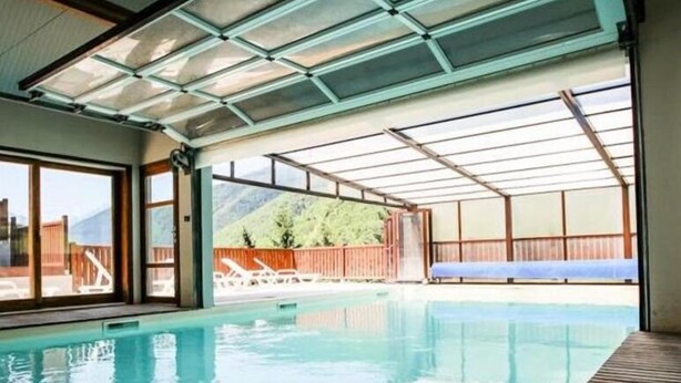 Una piscina interior con paredes de vidrio transparente, terraza de madera y vistas a un paisaje montañoso.