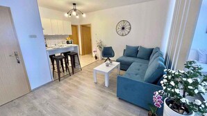 A modern living room with a blue sofa, a white coffee table, and a small kitchen area with a dining table and bar stools.