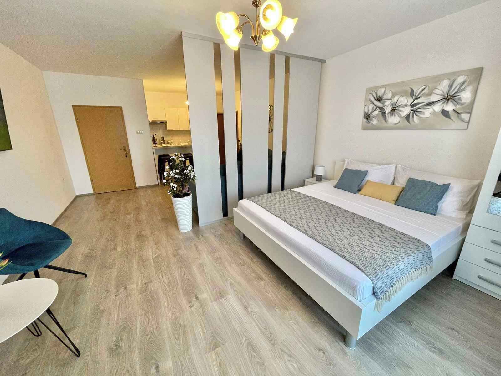 A modern bedroom with a large bed, a bedside table, a chair, a potted plant, and a kitchen in the background.