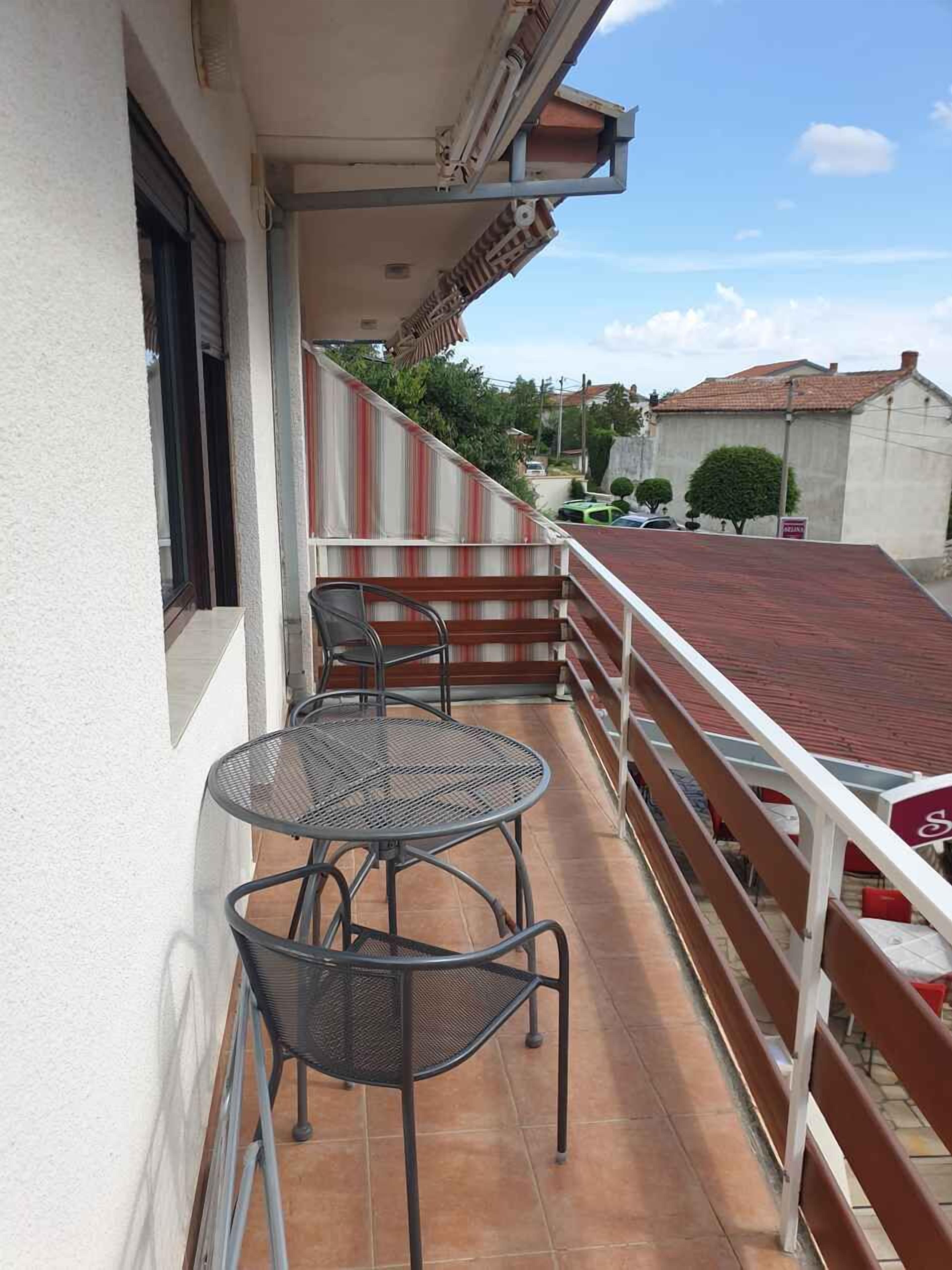 Un balcon avec une table en métal et deux chaises, un auvent rayé de rouge et de blanc, et une vue sur la rue et les immeubles.