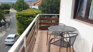 A balcony with a metal table and chairs, a view of a road, and a building with a window.