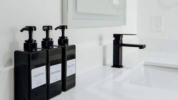 Een badkamer met een witte wastafel, drie zwarte shampoo flessen, een zwarte kraan en een grote spiegel.