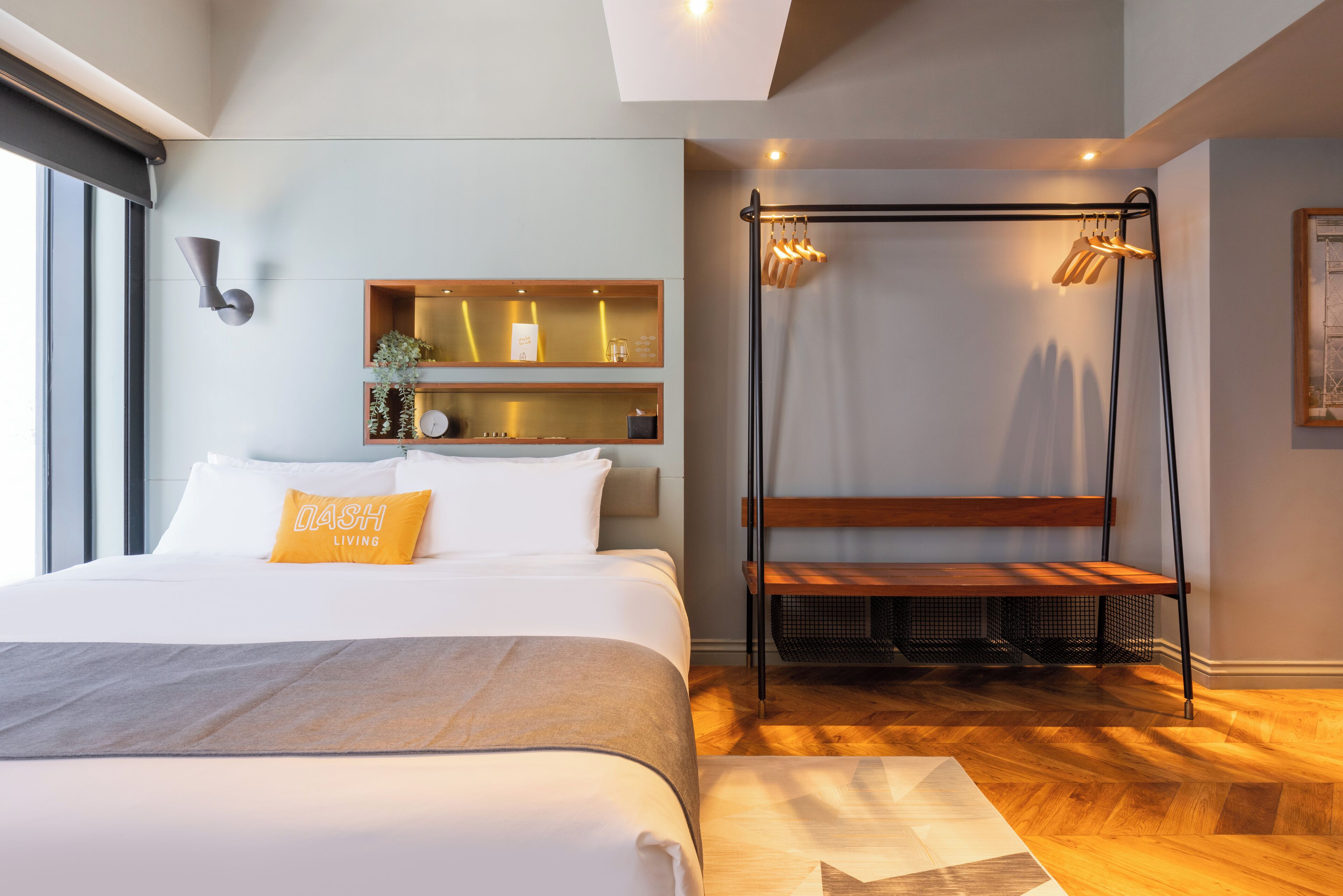 A modern hotel room with a large bed, a bench, a hanging light fixture, and a wall-mounted shelf with decorative items.