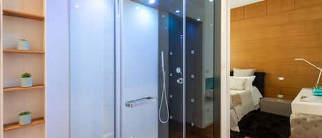 Une salle de bain moderne avec une douche à parois de verre, des étagères en bois ornées de plantes en pot, une table de chevet avec une lampe et un lit surmonté d’un tapis foncé.