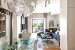 A modern living room with a glass dining table, a sofa, and a large window with a view of greenery. - Heima Smart - Charme Apartment (Palermo)
