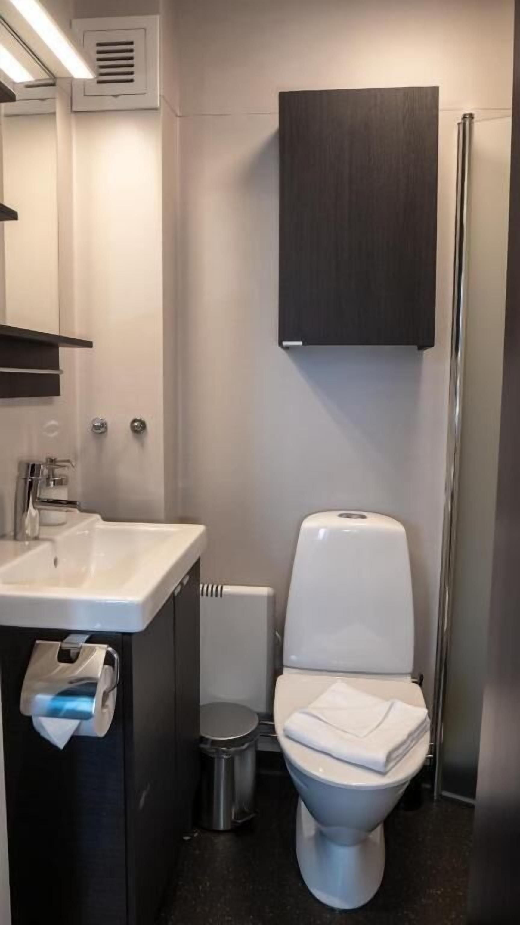 Une salle de bain moderne avec des toilettes blanches, un lavabo équipé d’un distributeur de serviettes en papier, un miroir et un meuble en bois foncé placé au-dessus des toilettes.
