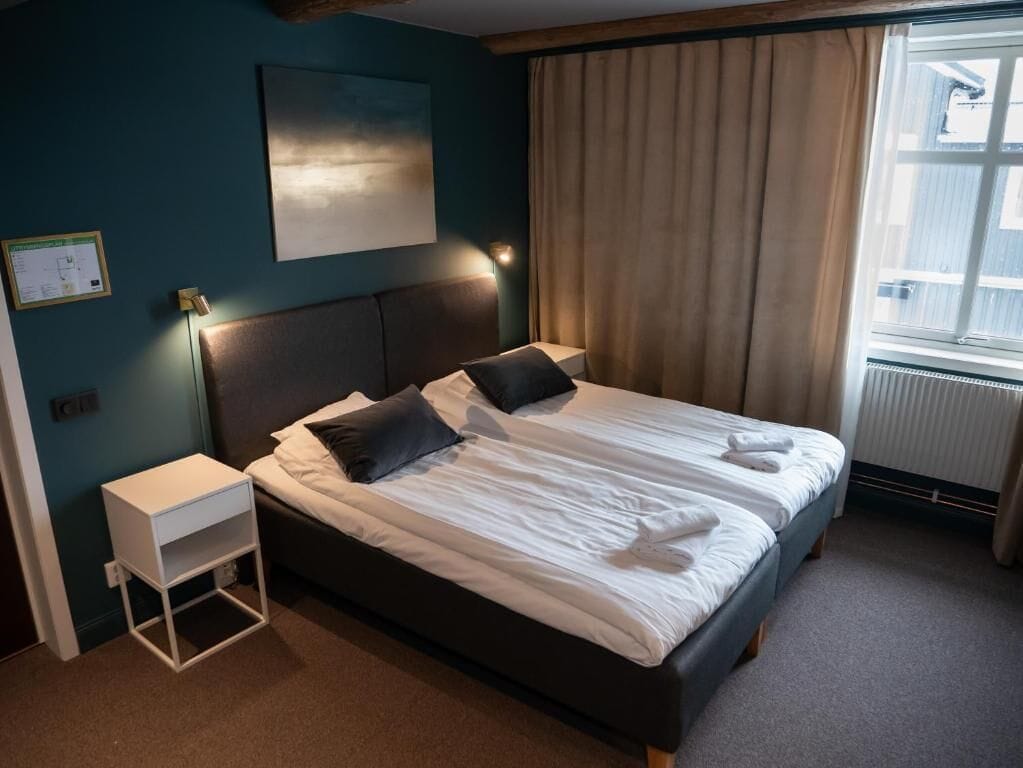 A neatly made bed with white linens and two pillows, a bedside table with a lamp, a wall-mounted artwork, and a window with curtains.
