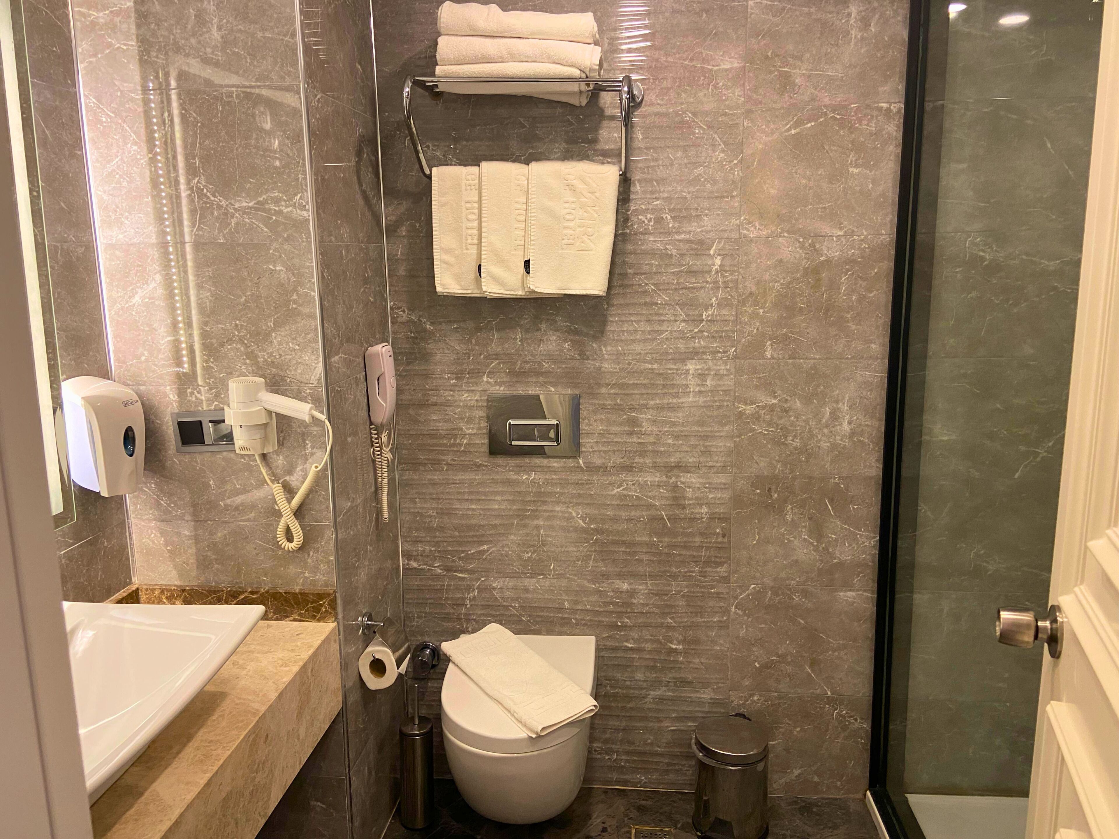 Une salle de bain équipée de toilettes, d’un sèche-mains, d’un porte-serviettes, d’une poubelle et d’une cabine de douche en verre.