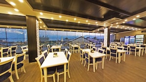 A restaurant with tables and chairs arranged indoors, overlooking a scenic view of a body of water and mountains. - Mara Palas Hotel (Fethiye)