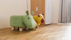 Dos peluches sobre un suelo de madera, uno verde y uno amarillo, con una pared blanca y una puerta al fondo.