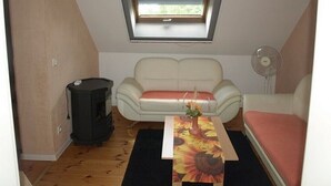 A small, cozy room with a white sofa, a table with a sunflower tablecloth, a heater, and a window with a view.
