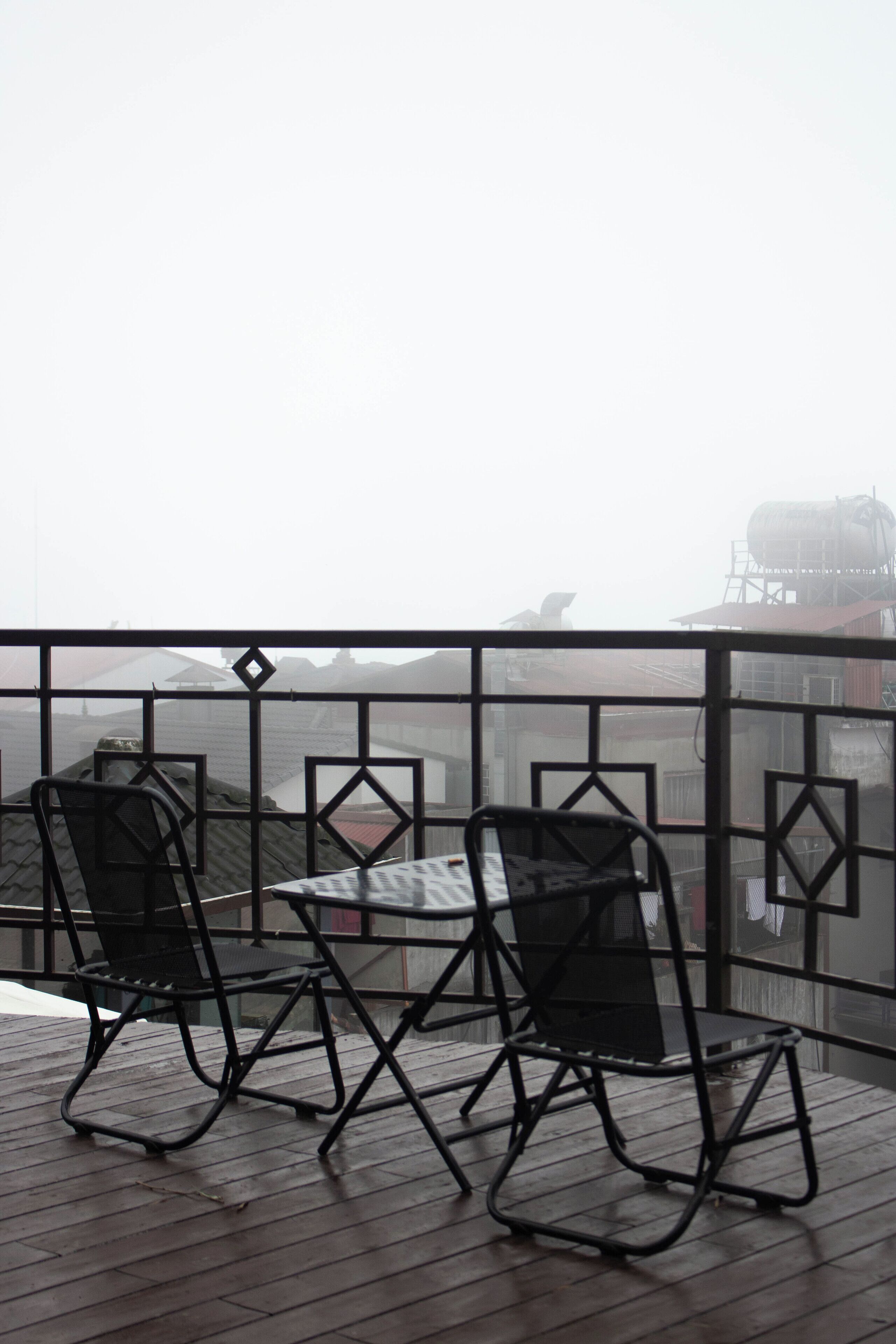 Un balcon avec deux chaises en métal noir et une table, donnant sur un paysage urbain embrumé.