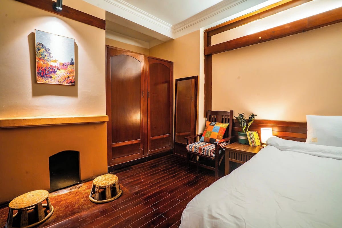 A hotel room with a bed, wooden wardrobe, a chair with a colorful cushion, a small stool, and a painting on the wall.