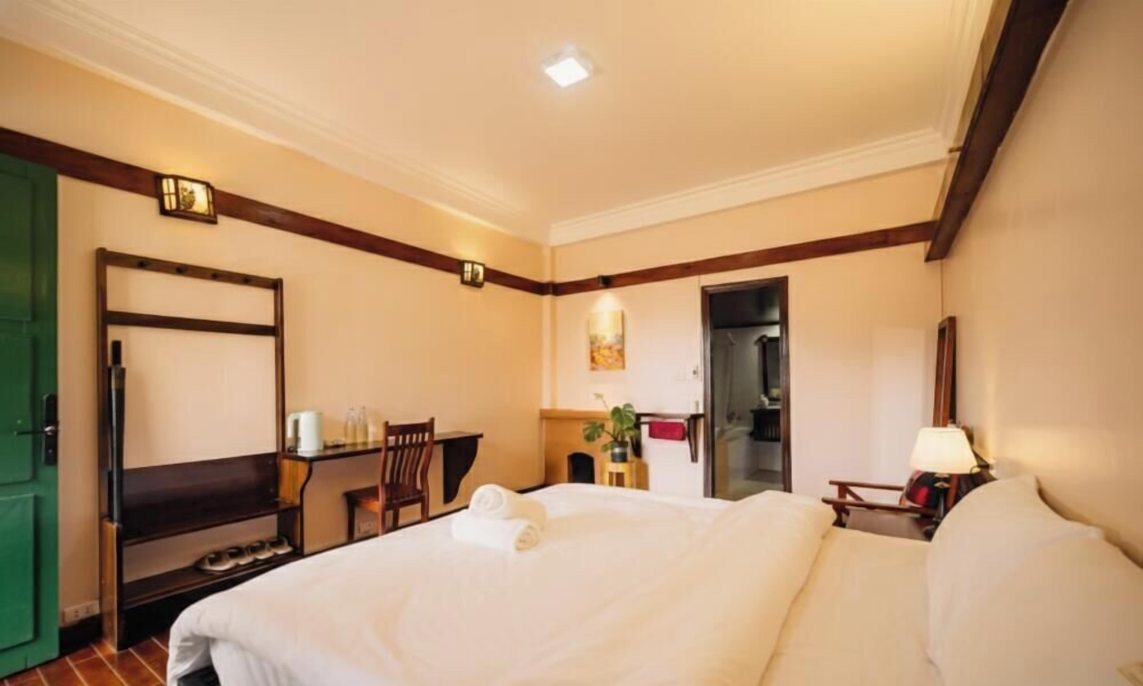 A hotel room with a bed, a desk, a chair, a lamp, and a green door.