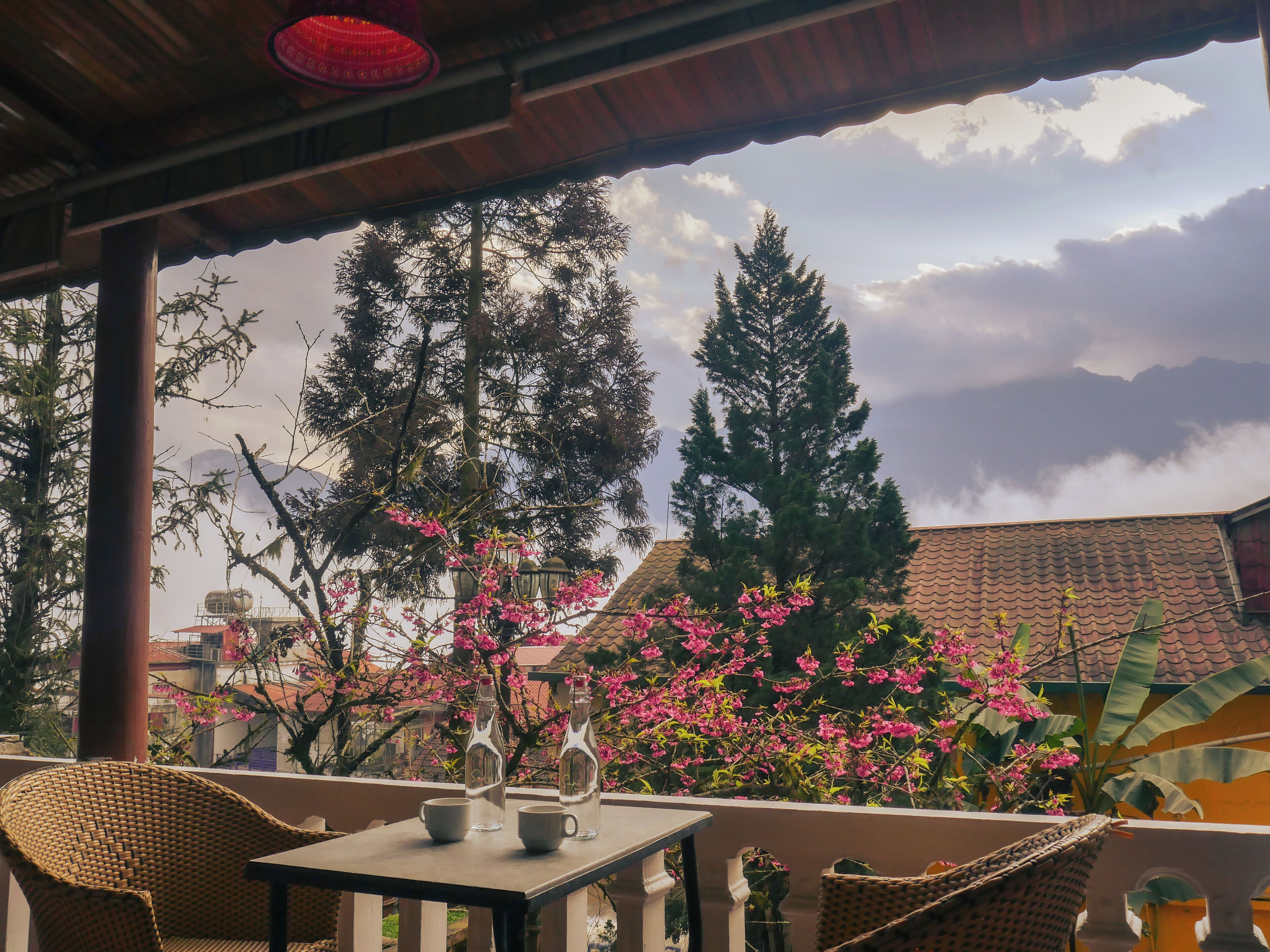 A covered outdoor seating area with wicker furniture, a table with two bottles and a cup, and a view of trees and buildings.