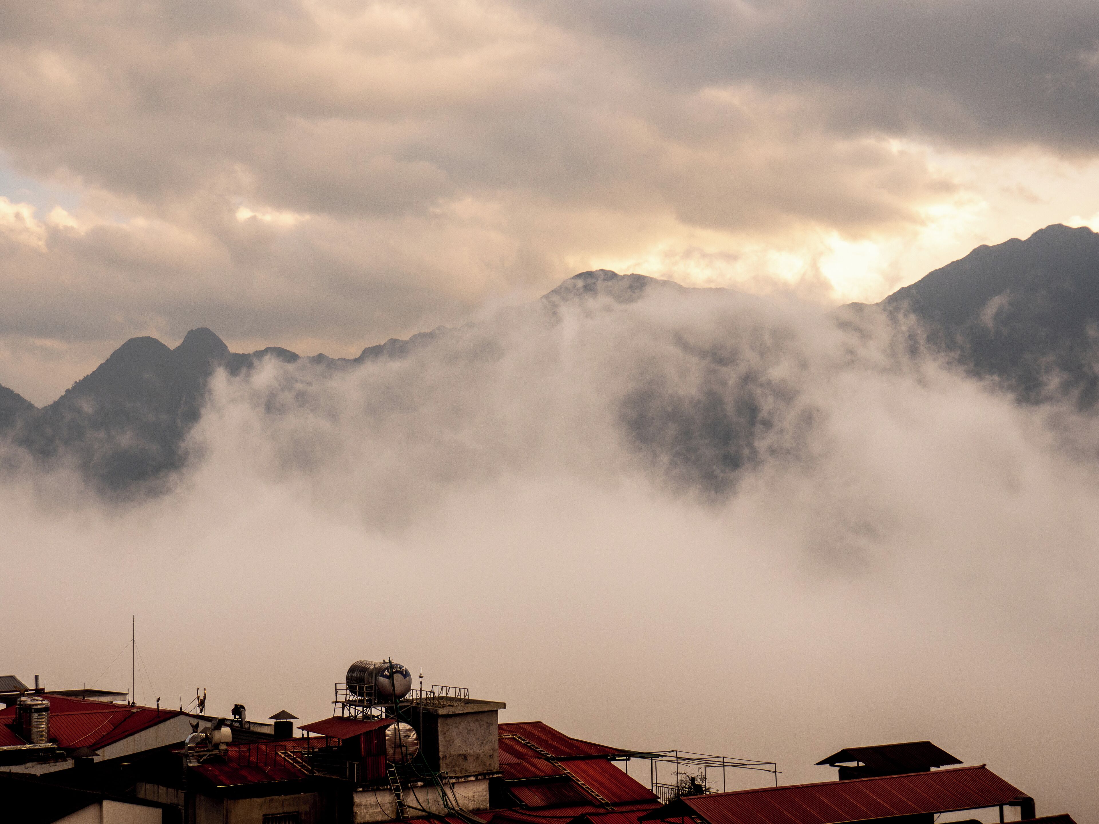 Una cordillera con nubes en la base y edificios con tejados rojos en primer plano.
