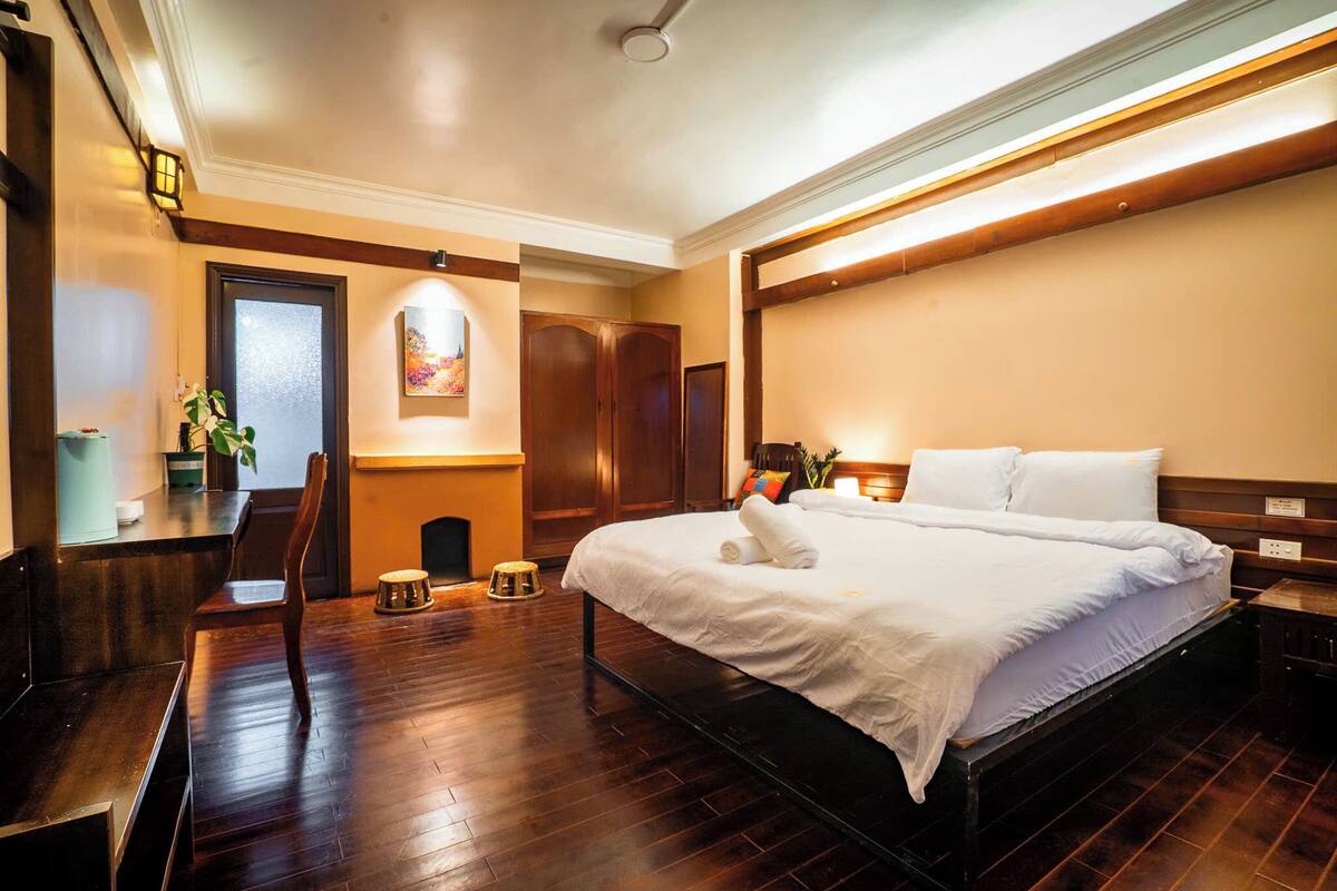 A hotel room with a large bed, wooden flooring, a desk with a chair, and a small stool.