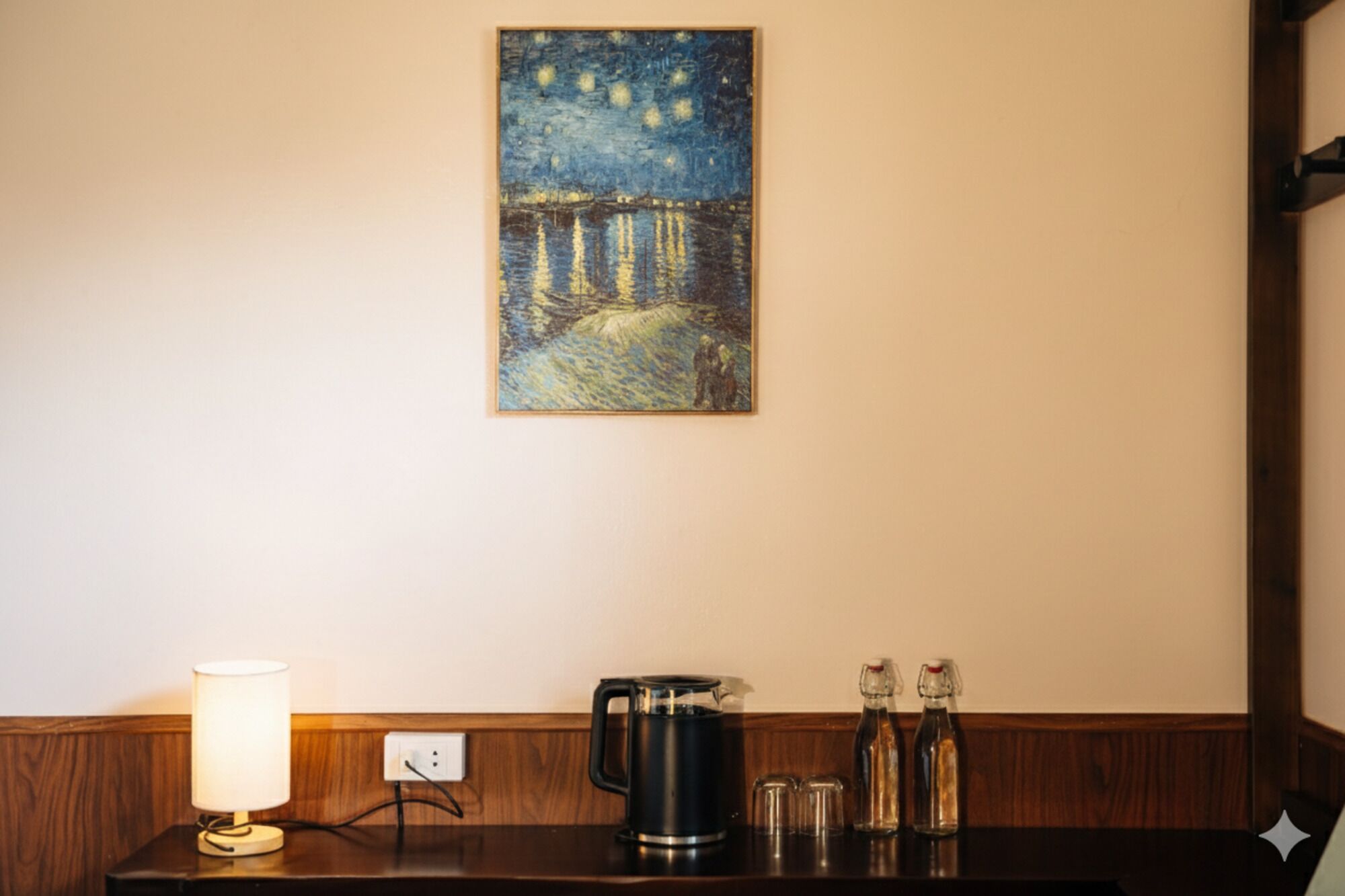 Habitación de hotel con un cuadro de una escena nocturna, una mesita de noche con una lámpara, una cafetera y saleros y pimenteros.