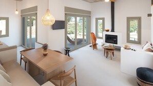 A modern living space with a wooden dining table, a sofa, a TV mounted on the wall, and a fireplace. - Themis Homes Thallo  in Skyros (Skyros)