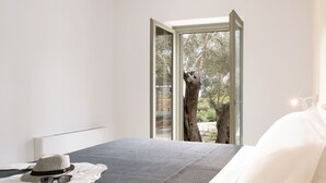 A modern bedroom with a large bed, a view of trees through a glass door, and a hat and sunglasses on the bed. - Themis Homes Thallo  in Skyros (Skyros)