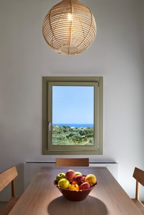 Een eetgedeelte met een tafel, een fruitschaal, een rieten hanglamp en uitzicht op zee door een raam.