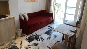 A compact living space with a red sofa, a small table, and a kitchen area with a stove. - Castelopetra Studios (Amorgos)