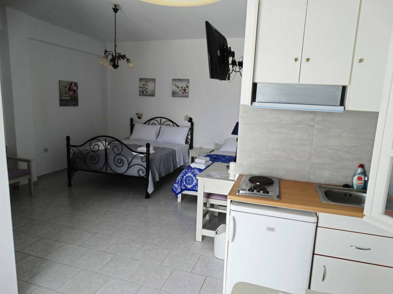 A bedroom with a bed, a bedside table, a small desk, and a kitchenette area with a stove and cabinets.