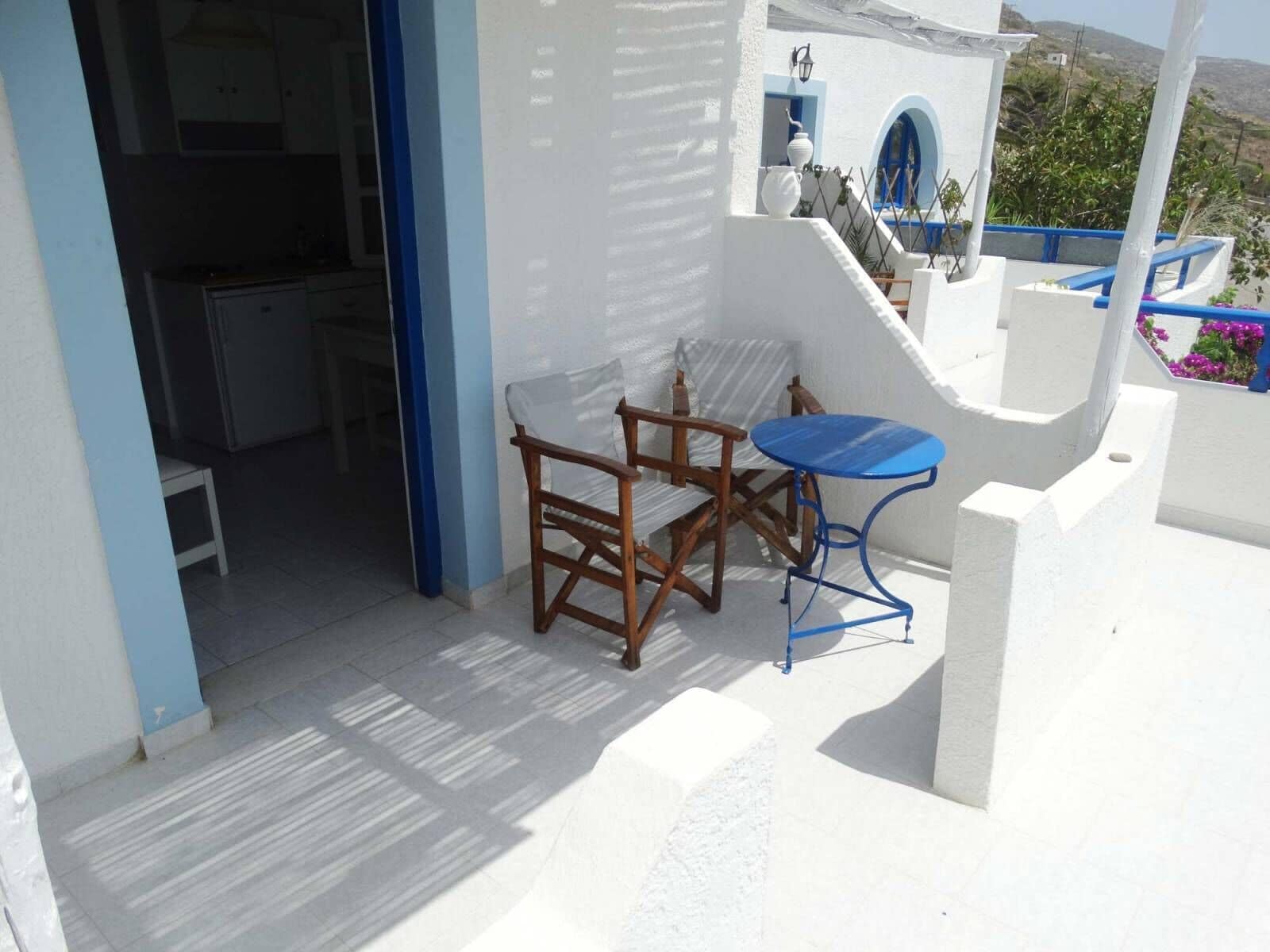A balcony with a table and chairs, a view of a building with blue accents, and a door leading to an interior space.