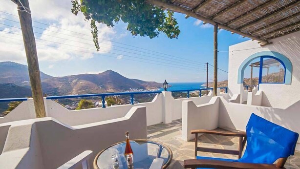 A terrace with a glass table, blue chairs, and a view of the sea and mountains. - Castelopetra Studios (Amorgos)