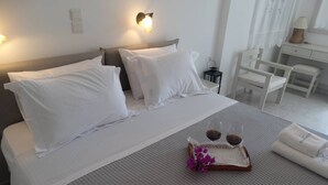 A neatly made bed with white linens, a tray with two glasses of red wine, and a small bouquet of purple flowers. - Castelopetra Studios (Amorgos)