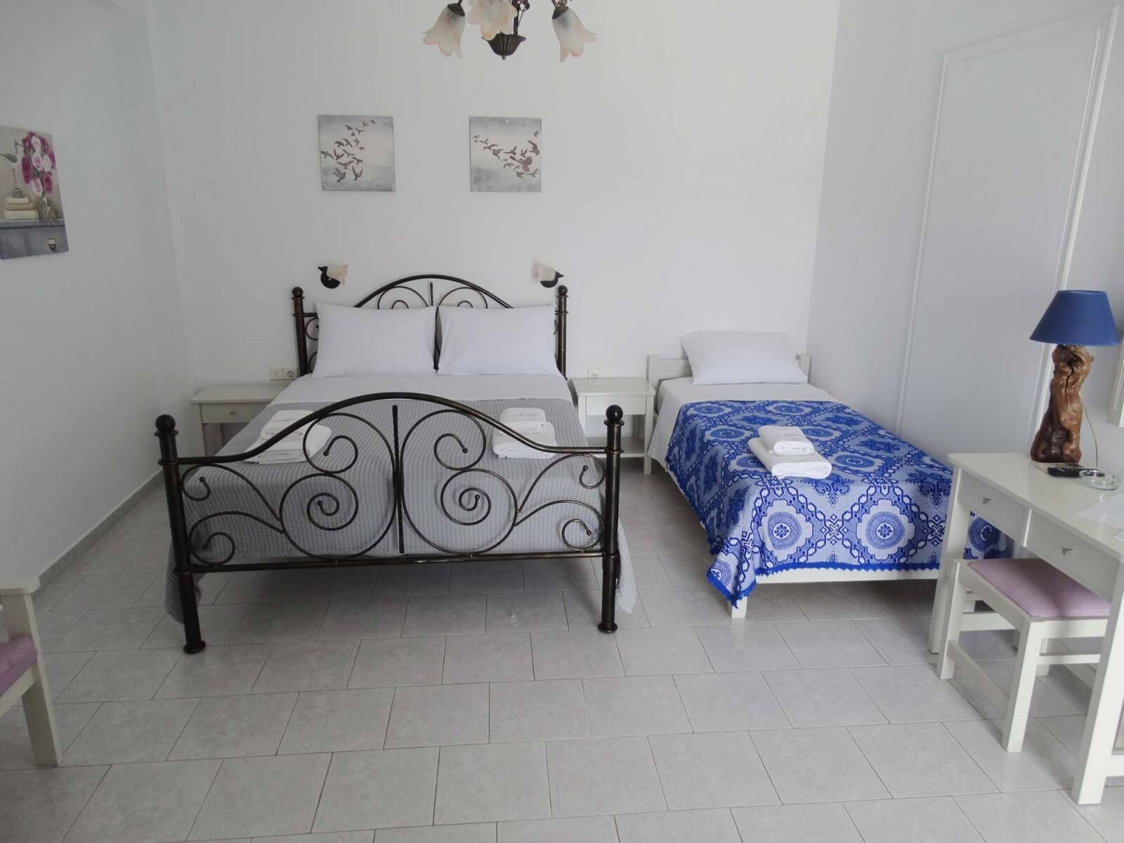 A bedroom with a large bed, a smaller bed, a desk, and a chair. There are two wall-mounted lamps and a painting on the wall.