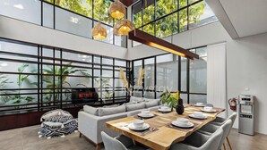 A modern dining area with a wooden table set for four, surrounded by grey upholstered chairs. A grey sofa and a black and white patterned pouf are visible in the background. - Elio Villa by Kozystay (Jimbaran)