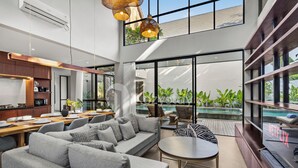 A modern living area with a sofa, dining table, and a TV. There is a large window with a view of a pool and greenery. - Elio Villa by Kozystay (Jimbaran)