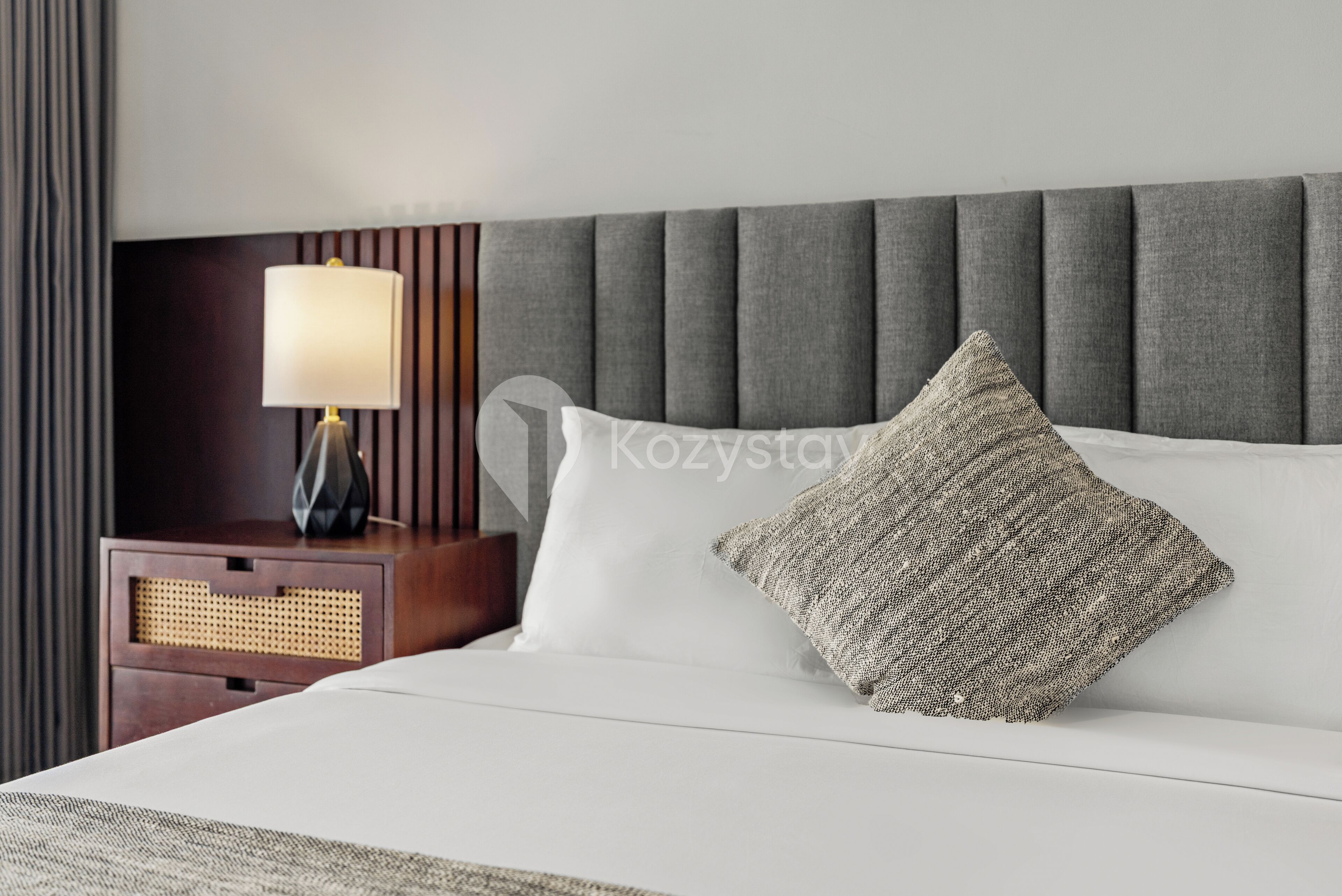 Una cama bien hecha con un cabecero tapizado gris, una lámpara de noche y una almohada con textura.