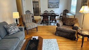 Un salon avec un canapé gris, des fauteuils à motifs, une table à manger avec des chaises, un coin cuisine et un tableau au mur.