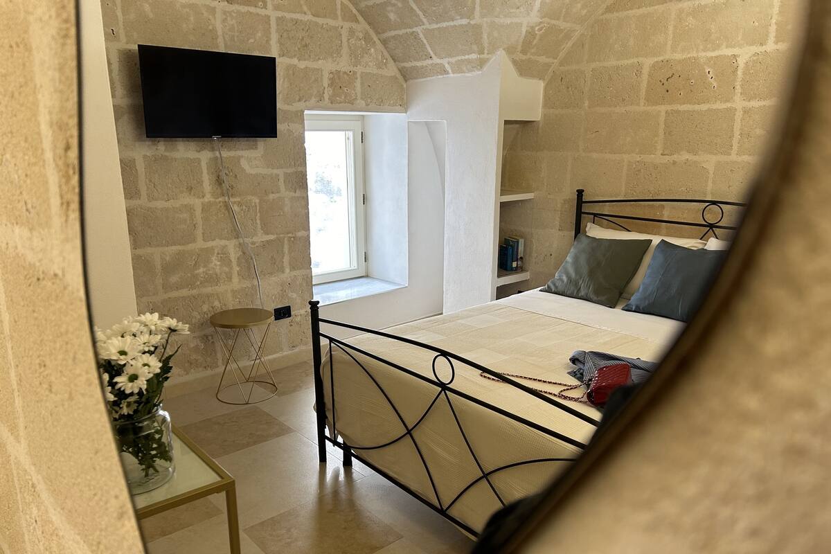 Une chambre avec un mur en pierre, un lit avec un cadre en métal, une table de chevet ornée d’un vase de fleurs, un téléviseur fixé au mur et une fenêtre donnant sur l’extérieur.
