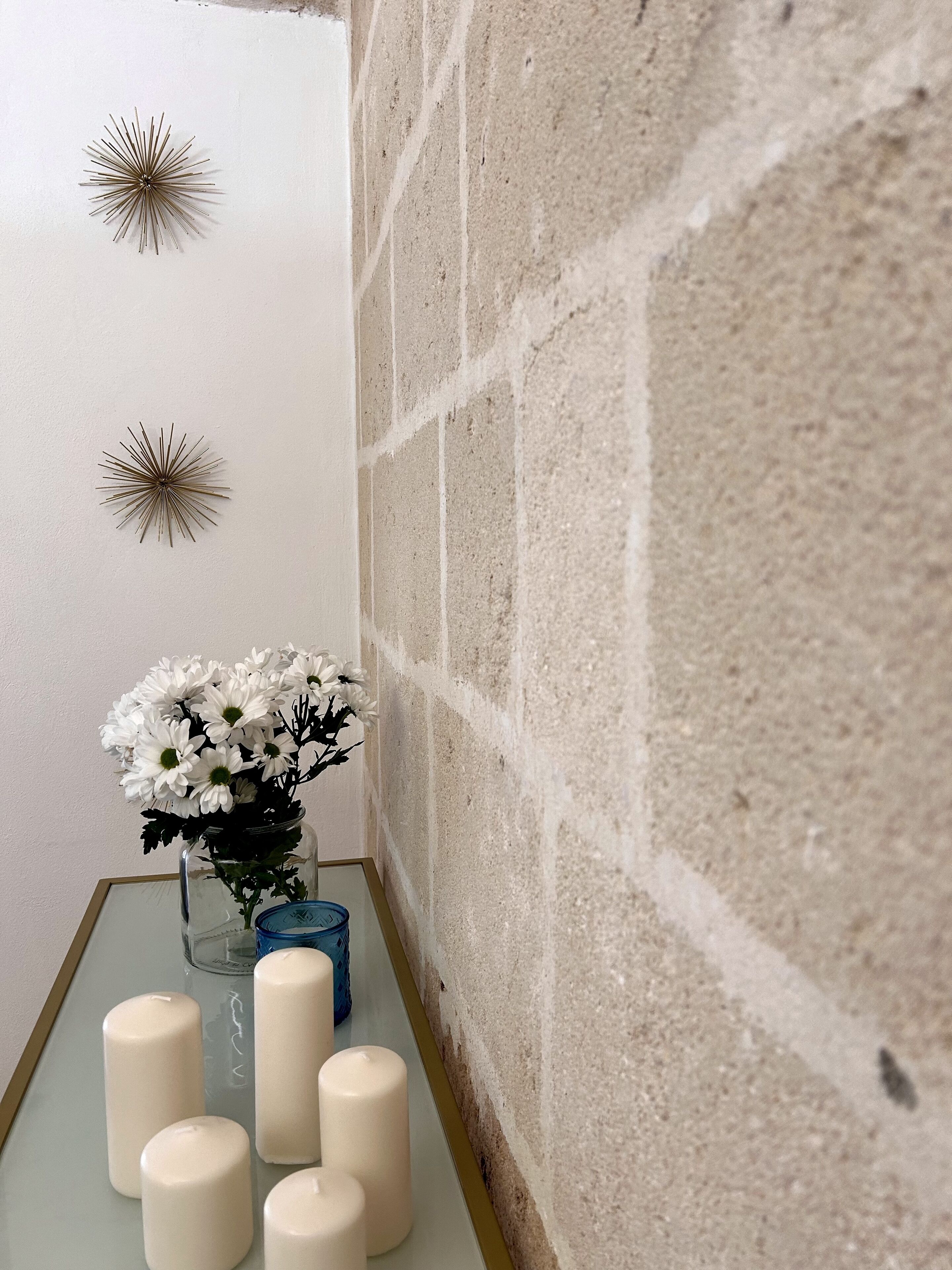Un coin de pièce avec un mur blanc, un mur en pierre à la texture particulière, une table en verre sur laquelle est posé un vase rempli de fleurs blanches, et plusieurs bougies blanches.