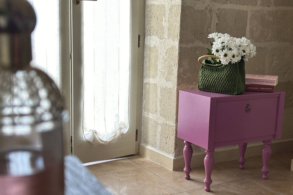 Une pièce avec une table d’appoint violette sur laquelle repose un panier tressé contenant des fleurs blanches, une porte ornée d’un rideau léger et un mur en pierre.
