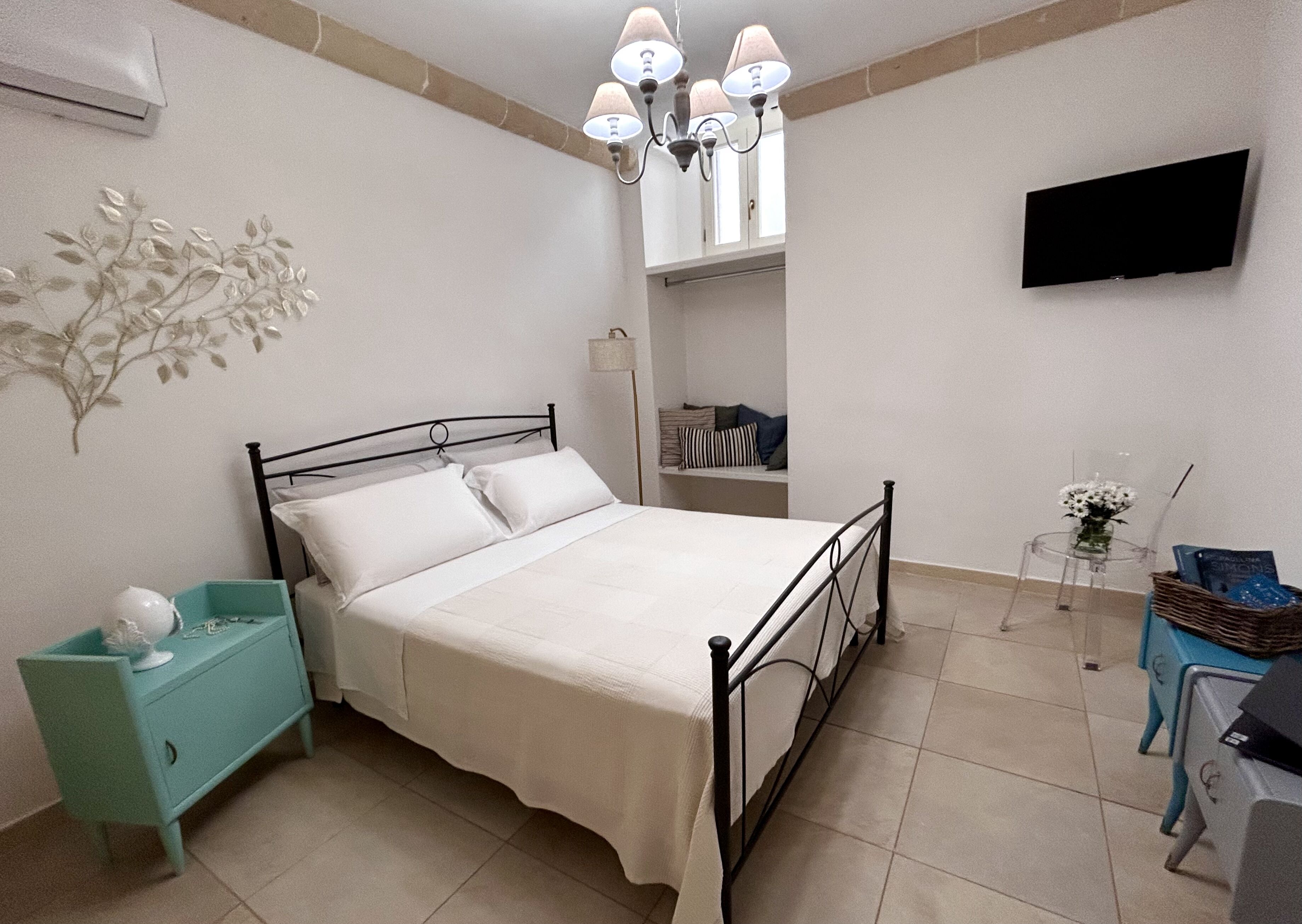 A bedroom with a bed, a nightstand, a dresser, a TV, a chandelier, and a window.