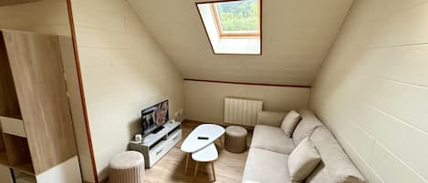 A small, cozy room with a sloped ceiling, a TV, a sofa, two ottomans, and a radiator.